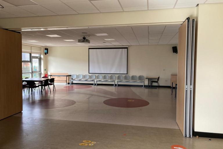 Spacious room with three available tables and several chairs placed close to the walls. Two folding doors that allow for a spacious entrance to the room and the wall is composed of windows. In the middle of the back wall, opposite to the entrance, there is a large projector screen. 