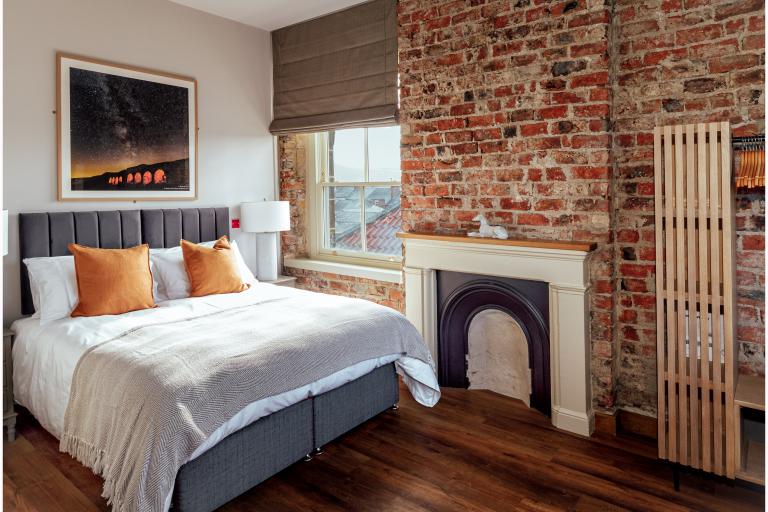 One of the bedrooms from the Guisborough Town Hall. The image shows a double bed placed next to a window, a brick wall and a fire place.