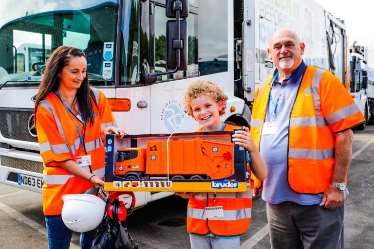 Image of a young boy and his mother holding a bin wagon toy offered by Cllr Barry Hunt on behalf of the council. Behind them there is a real bin wagon.