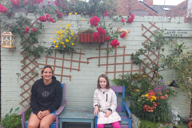 Image of a woman and her daughter sat on a bench in their colourful alleyway with flowers behind and around them.