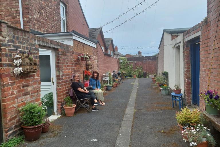 Image of two women sat on chairs in their alleyway which is decorated with flowers and outside furniture. 