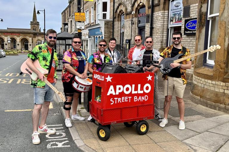 A group of musicians dressed colourfully and holding guitars gathered around a red cart holding big speakers. 