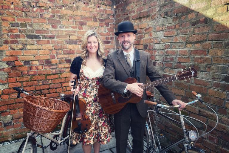 A woman dressed in a flower patterned dress and a man wearing a suit and a hat next to two vintage bikes. 