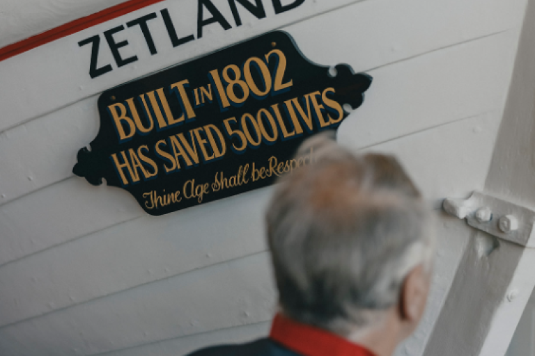 Picture from the RNLI exhibition featuring the tip of the white Zetland boat. The boat wears a plaque stating that the boat was built in 1802 and saved 500 lives.