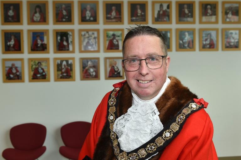 Image of Neil Bendelow, who has been elected as mayor. Neil is wearing a red ceremonial jacket with gold and silver jewellery around his neck. In the background there are images of the previous mayors.