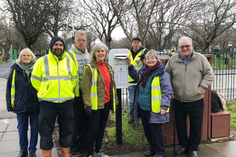 Image of volunteers standing in front a white postbox. They are all wearing yellow hi vis jackets.