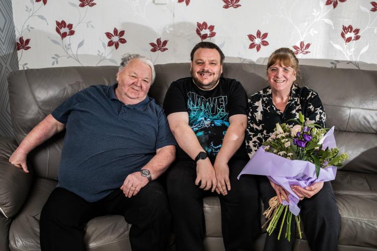 Two man and a woman sat on a couch, the woman holding a bouquet of flowers.