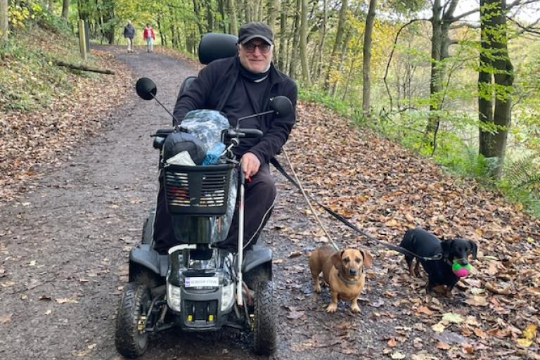 A man on a disability scooter holding to small dogs on a leash on a forest path.