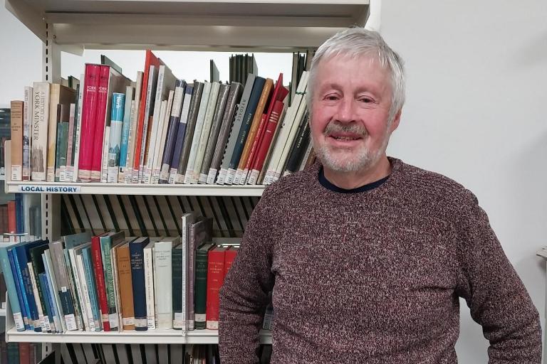 a man dressed in a brown shirt standing next to a bookshelf full of books of different colours