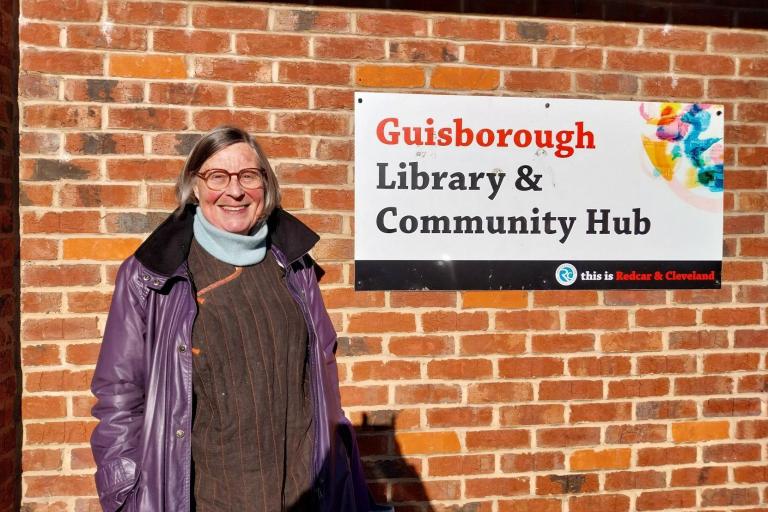 A woman standing next to a sign that reads Guisborough Library & Community Hub. Behind her there is a brick wall.