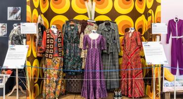 Dresses from various decades in history exhibited. A purple dress in the middle, a grey suite on the right hand side and an orange one on the far right. On the left hand side two multicoloured dresses.