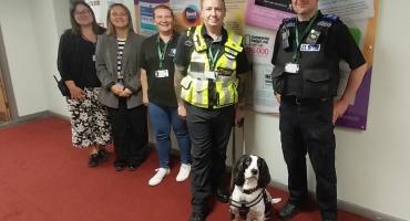 Image of two police officers and three council officers standing in a school hallway with a leashed black and white dog.