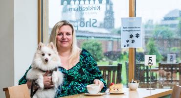 Image of a woman in a green dress sitting at a table and having tea while holding a small white dog. Behind her there is a sign reading: "Table reserved for our four-legged customers"."