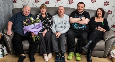 A group of five people, three men and two women, gathered on a grey couch and smiling