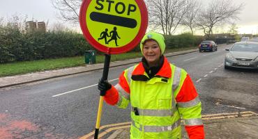 Image of female lolipop lady. She is wearing a green and orange jacket holding a yellow and red sign that says the word 'stop' on it. 