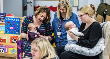 Image depicts three women, two of them holding babies, in a playroom.