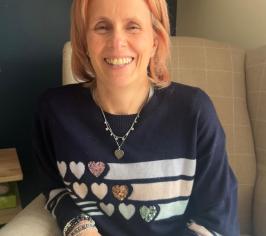 Image of a woman sitting on a light brown chair. She is wearing a blue shirt with multicoloured heart logos.