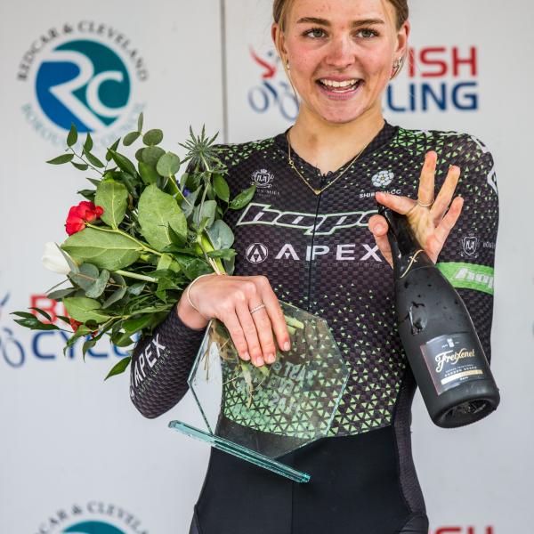 Winner of the women's race wearing her cycling gear, holding a bouquet of flowers and waving t the camera. 